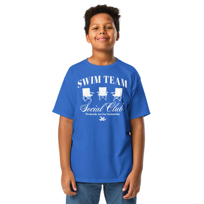 Boy wearing royal blue Swim Team Social Club Youth T-shirt by TrendySwimmer, featuring foldable chair graphic and swim lifestyle slogan.